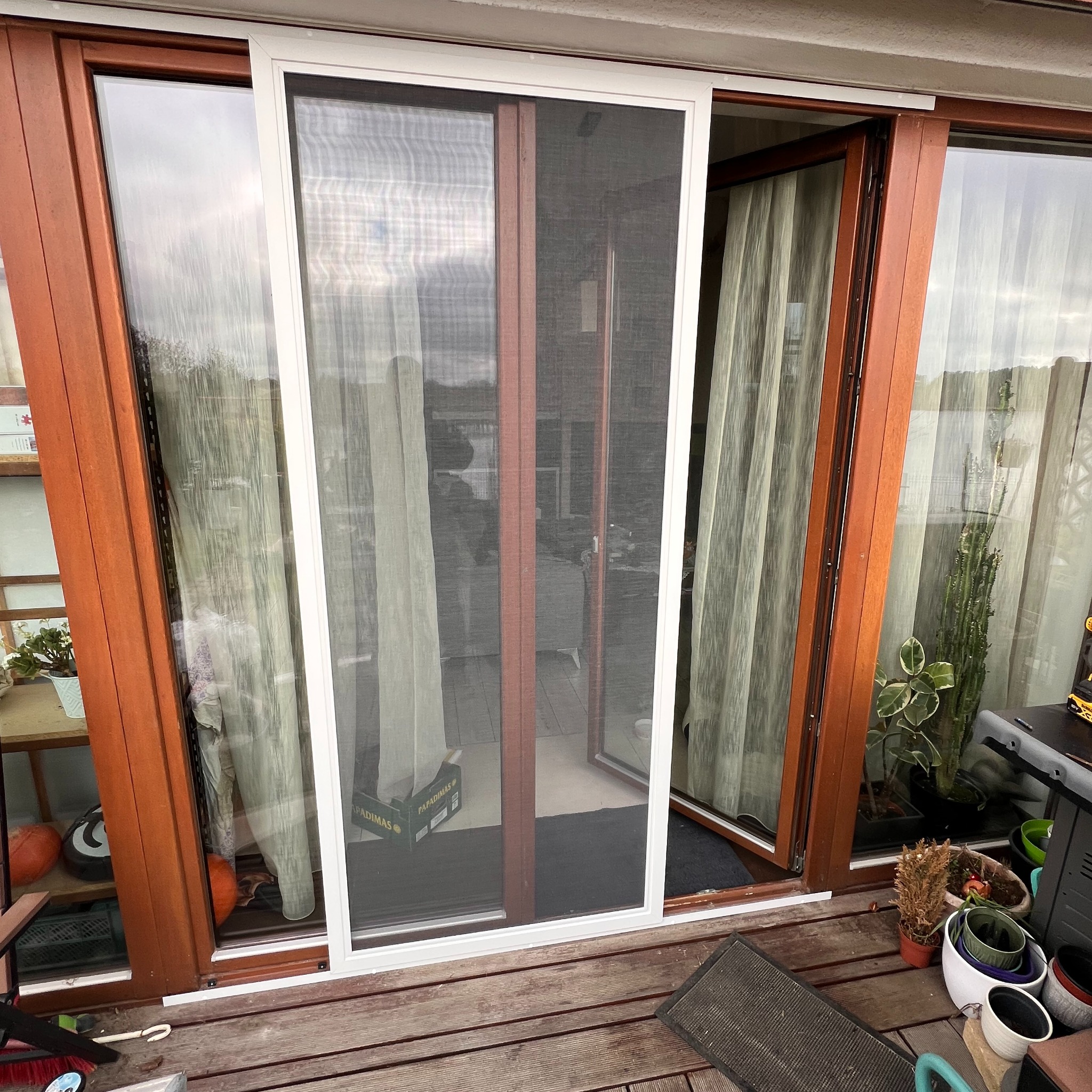 Zamontowana przesuwna moskitiera w białej ramie na drewnianym tarasie, widok z zewnątrz. Widoczne rośliny w donicach i elementy wyposażenia tarasu.