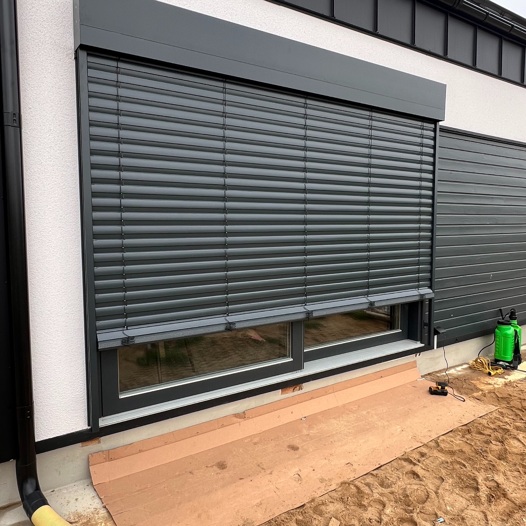 Grafitowa roleta zewnętrzna częściowo opuszczona na oknie budynku z szarą elewacją. Widoczne narzędzia i osłona podłoża.