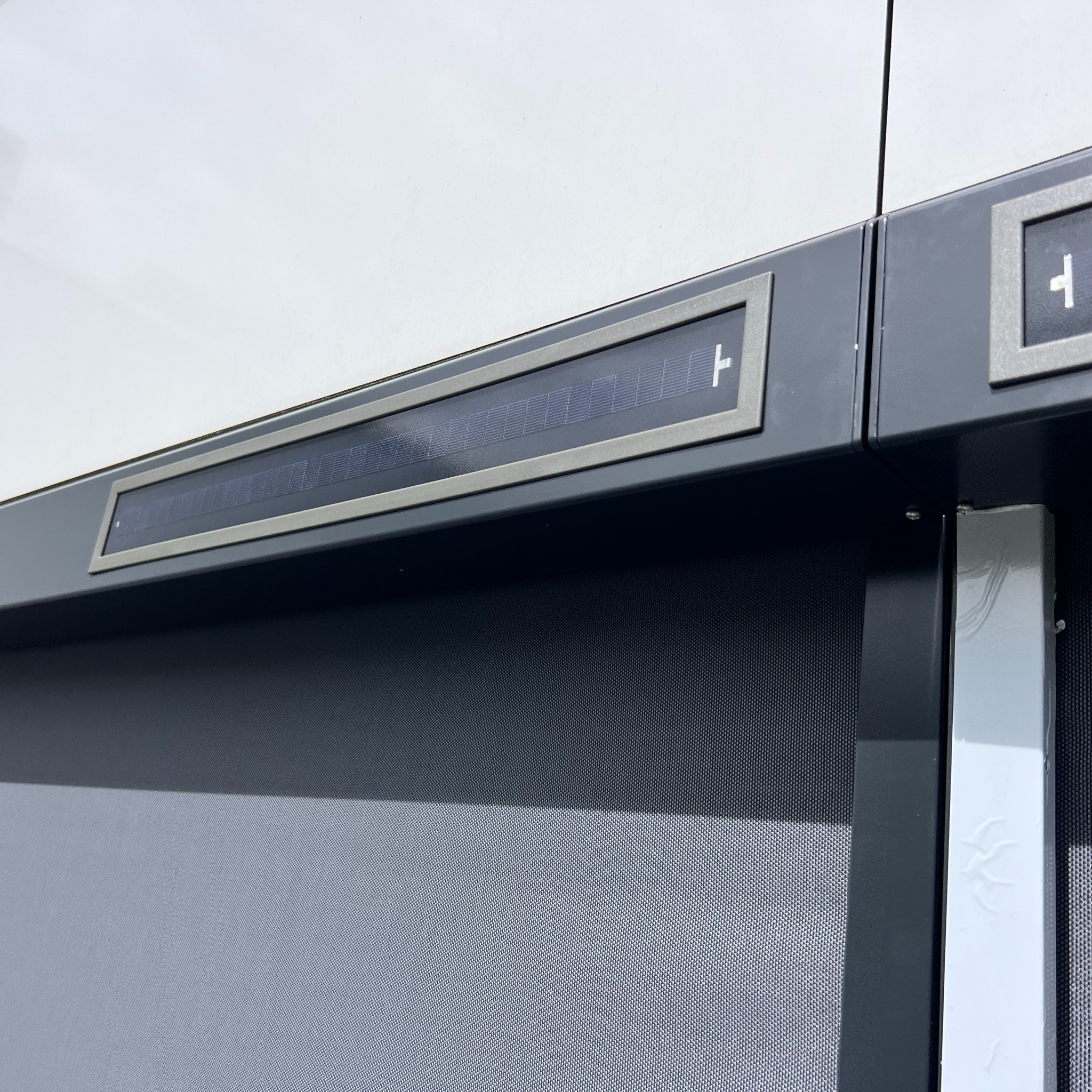 Szczegółowy widok zewnętrznej rolety z panelem solarnym w szarej ramie, zamontowanej na białej fasadzie budynku. Widoczna faktura siatki rolety.