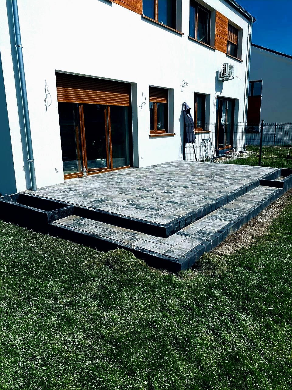 Nowoczesny taras z kostki brukowej w odcieniach szarości, z dwoma stopniami, przylegający do białego budynku z brązowymi oknami i zielonym trawnikiem.