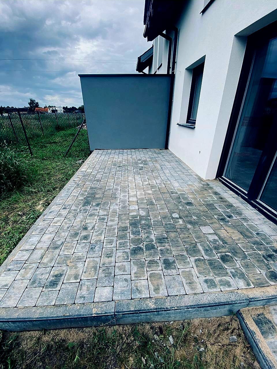 Nowoczesny taras z kostki brukowej w Kórniku. Szara kostka ułożona w regularny wzór, przylegająca do białej ściany budynku z dużym oknem.