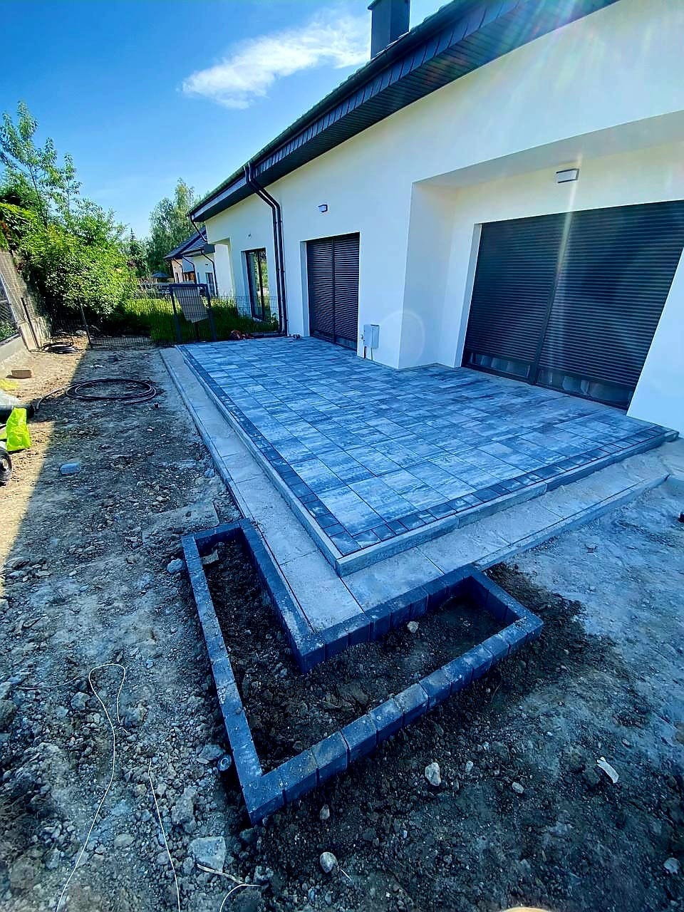 Nowoczesny taras z kostki brukowej w odcieniach szarości, z obrzeżami i przygotowanym miejscem na rabatę, na tle białego domu z ciemnymi bramami garażowymi.