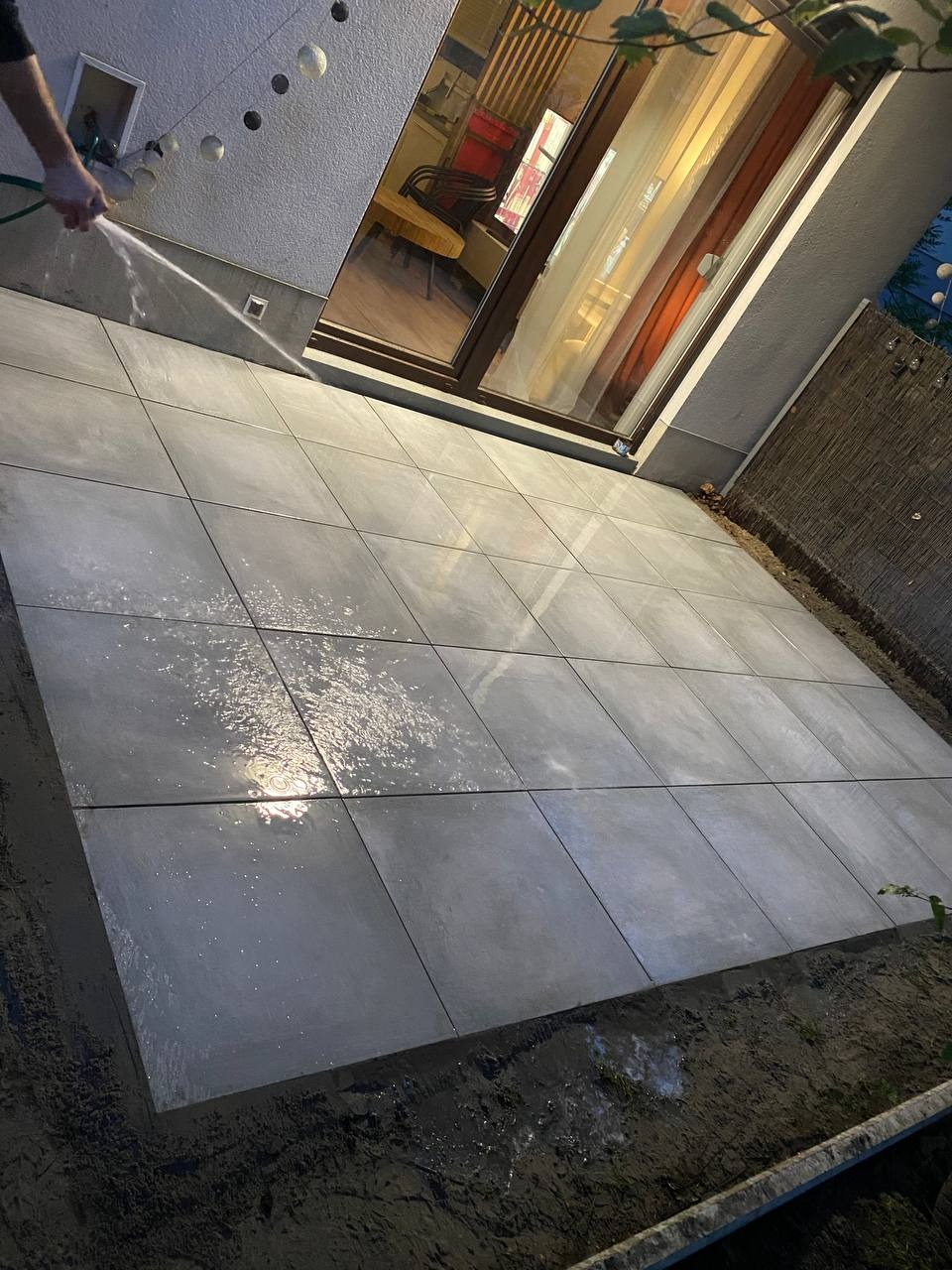Szare, kwadratowe płyty tarasowe, świeżo umyte wodą z węża, przylegające do ściany budynku z oknem tarasowym. Widoczne obrzeża i fragment ogrodu.