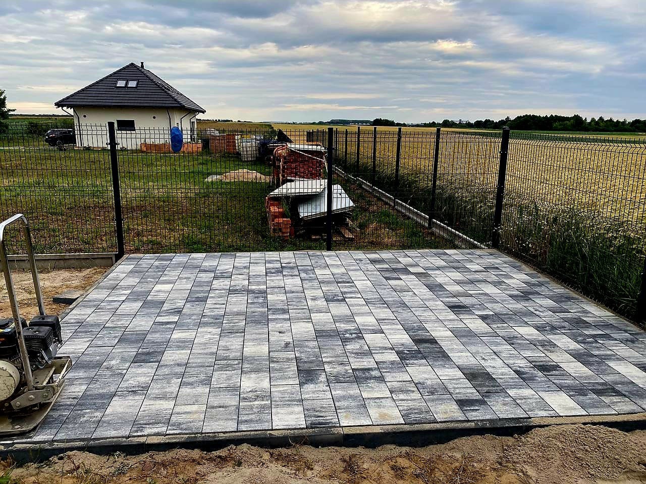 Ułożona, szara kostka brukowa na posesji z domem w tle, widok z góry. Częściowo widoczna zagęszczarka. Nowoczesny wzór kostki.