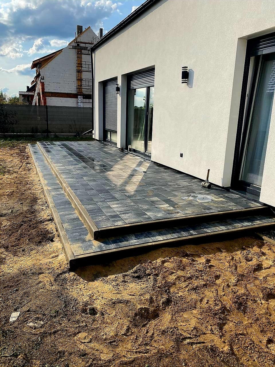 Nowoczesny taras z ciemnej kostki brukowej, dwupoziomowy, z widokiem na biały dom z dużymi oknami i budowę w tle. Widoczne niedokończone prace ziemne.