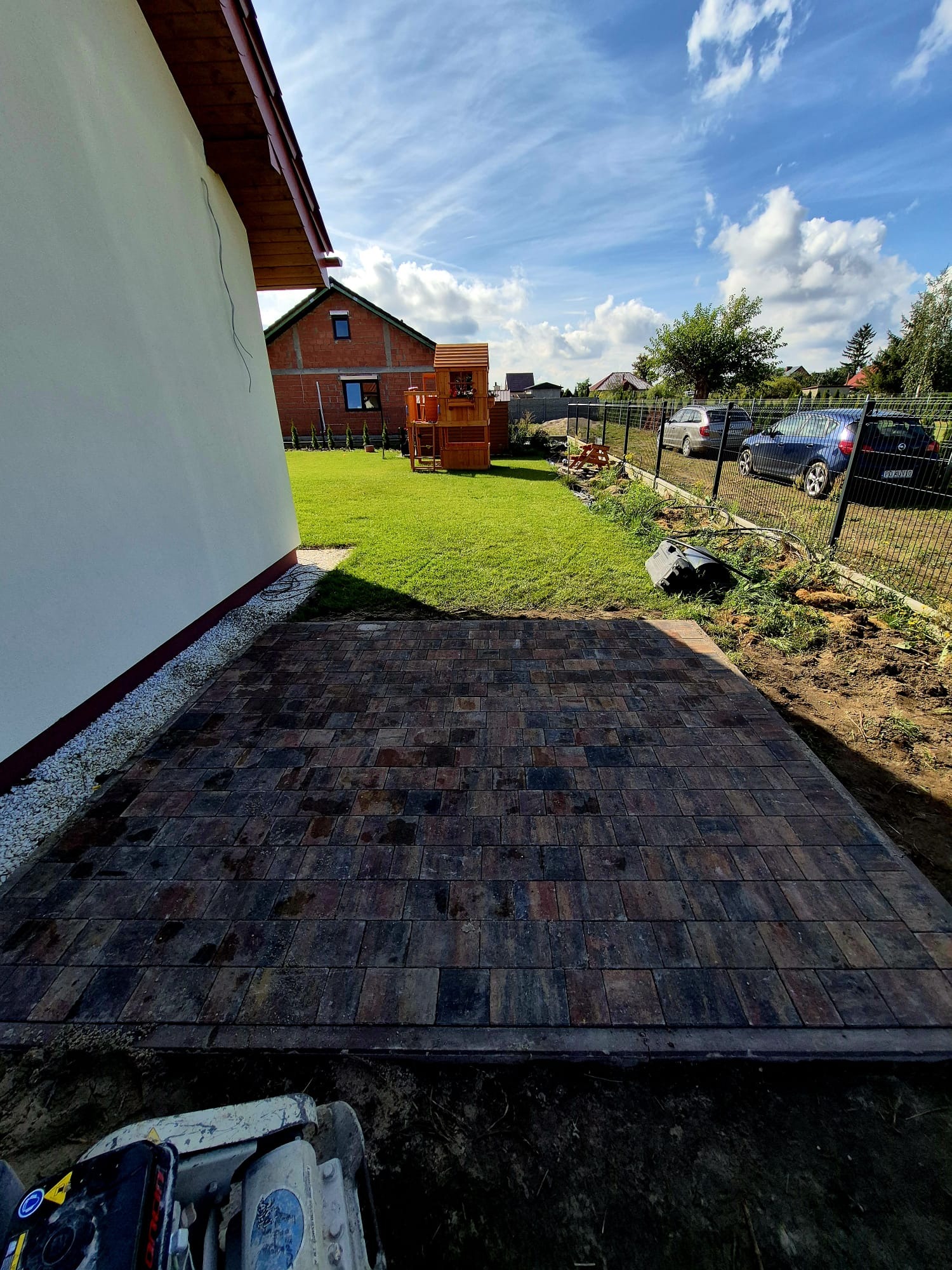 Ułożona kostka brukowa w odcieniach brązu i szarości, przylegająca do ściany budynku. W tle trawnik, plac zabaw i ogrodzenie z siatki.