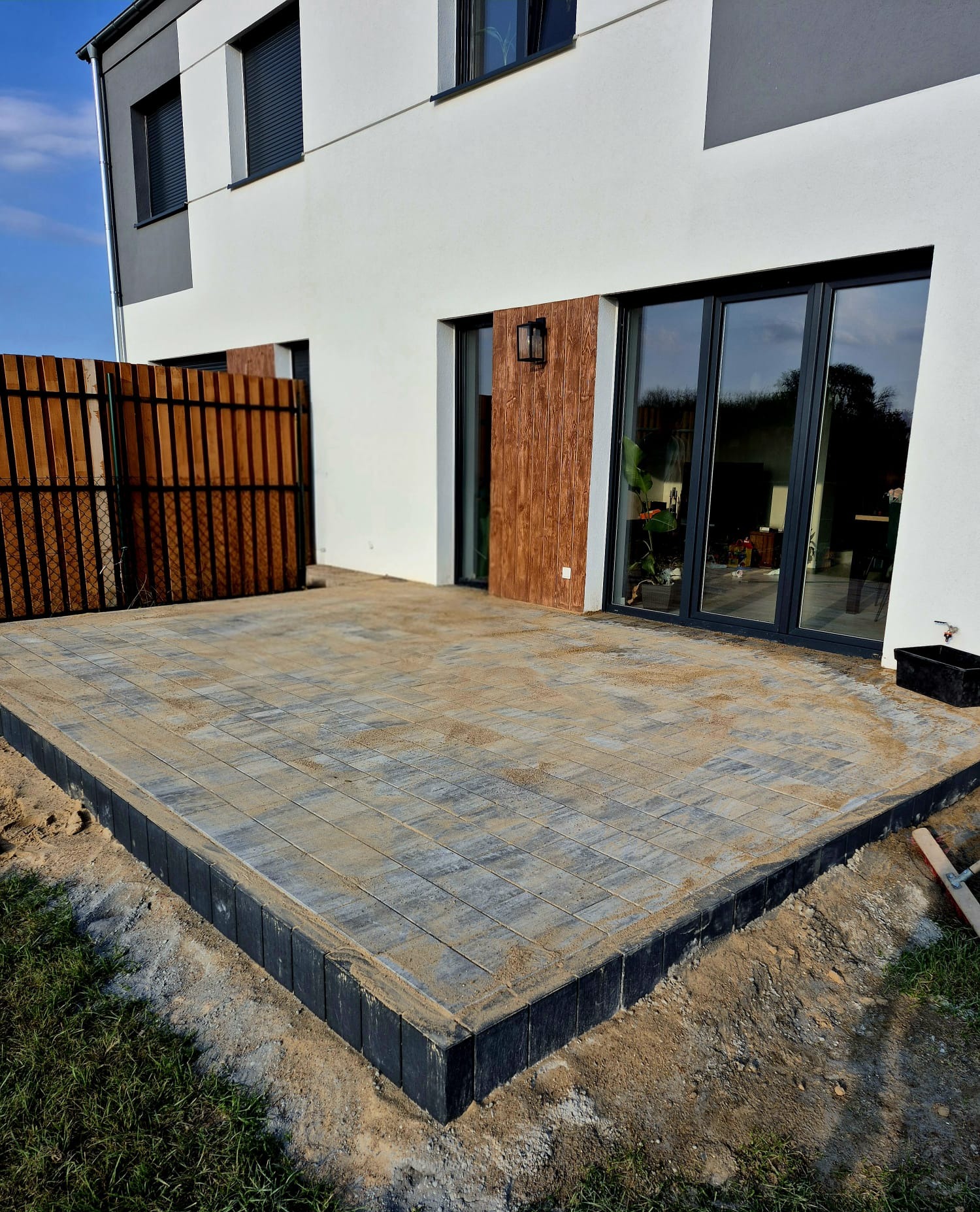 Nowoczesny taras z szarej kostki brukowej, obrzeża z ciemnych elementów, na tle białego domu z dużymi oknami i drewnianymi akcentami. Widok z góry, częściowo zasypany piaskiem.