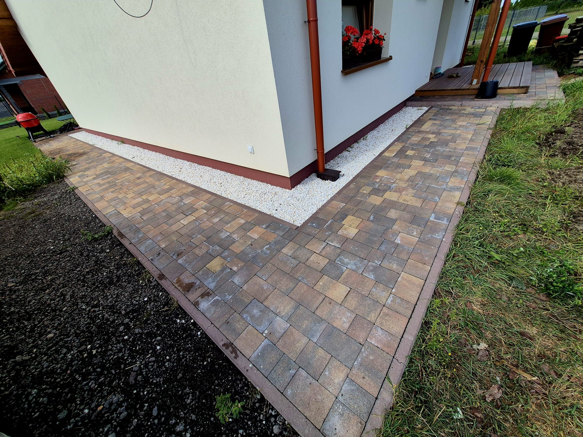Ścieżka z kostki brukowej w odcieniach brązu i beżu, obrzeża z czerwonej kostki, biały żwir przy ścianie domu, trawnik i rabata z czarną ziemią.