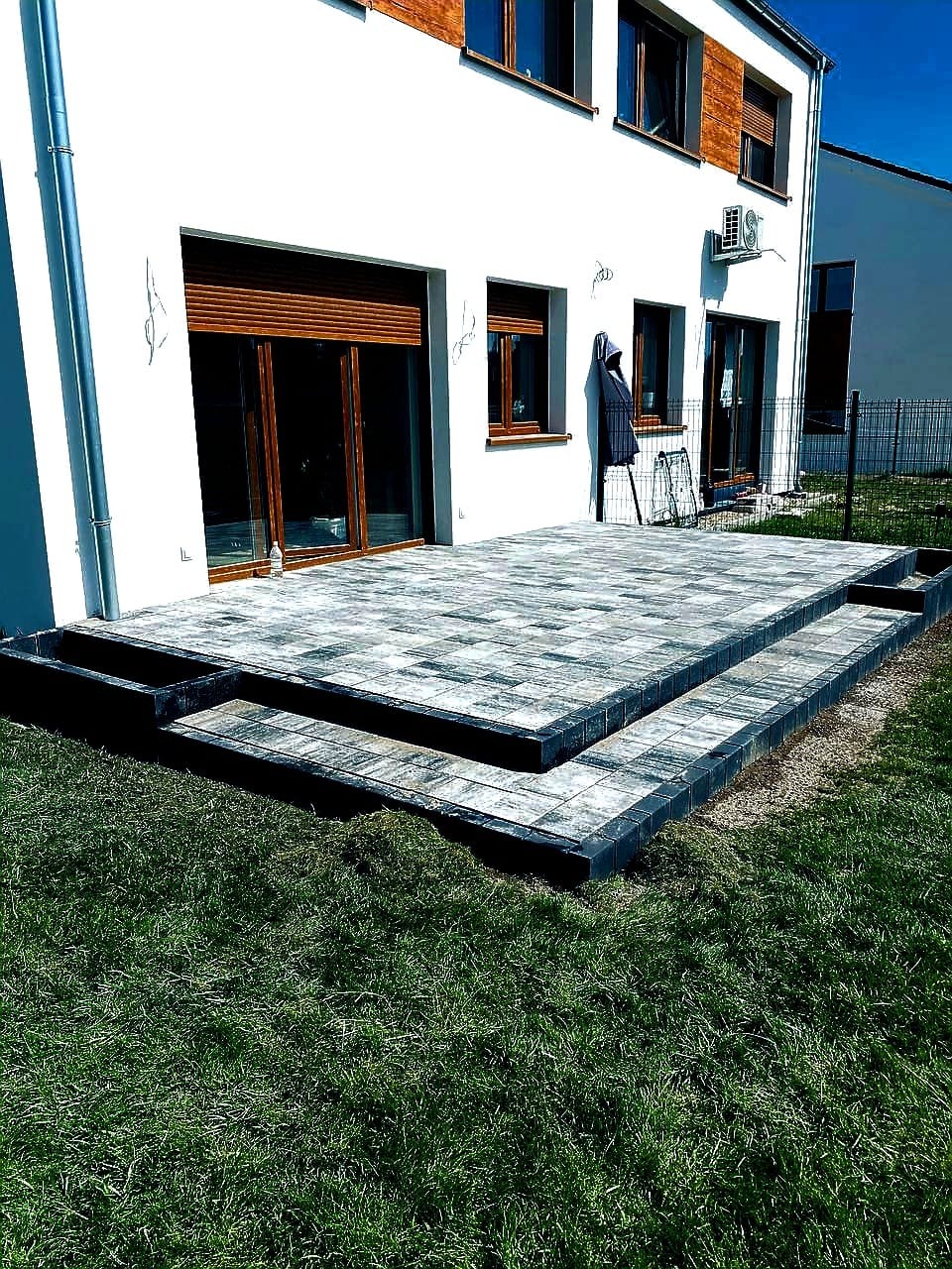 Wykończony taras z kostki brukowej w odcieniach szarości, z dwoma stopniami, przylegający do białego budynku z brązowymi okiennicami i zielonym trawnikiem.