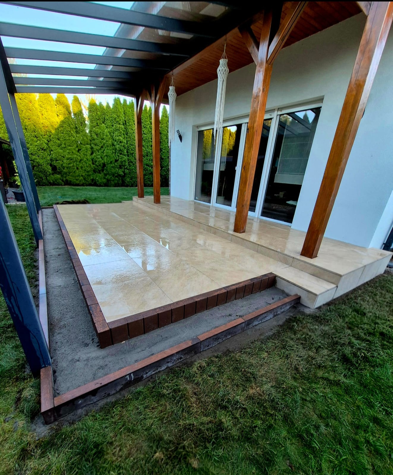 Wykończony taras z beżowych płytek ceramicznych, obrzeża z cegły klinkierowej, zadaszenie z drewnianymi słupami. Widok na ogród z tujami.