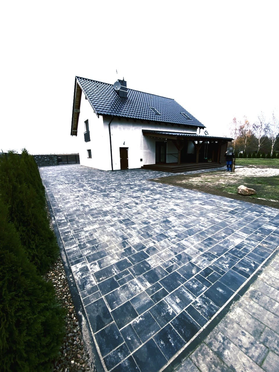 Ułożona, ciemna kostka brukowa prowadząca do białego domu z ciemnym dachem. Widoczny fragment ogrodu i osoba w tle. Ujęcie z niskiej perspektywy.