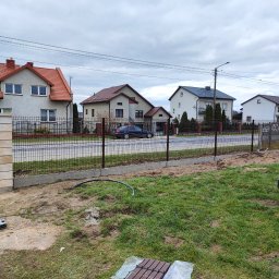Lukasz Ogrodzenia - Wznoszenie ogrodzenia panelowego z podmurówką i słupkami z imitacji piaskowca. Widoczne narzędzia i materiały budowlane. Tło stanowią domy jednorodzinne.