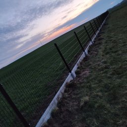 Lukasz Ogrodzenia - Długie, czarne ogrodzenie panelowe na betonowej podmurówce, rozciągające się wzdłuż zielonego pola pod wieczornym niebem z delikatnymi smugami różu.