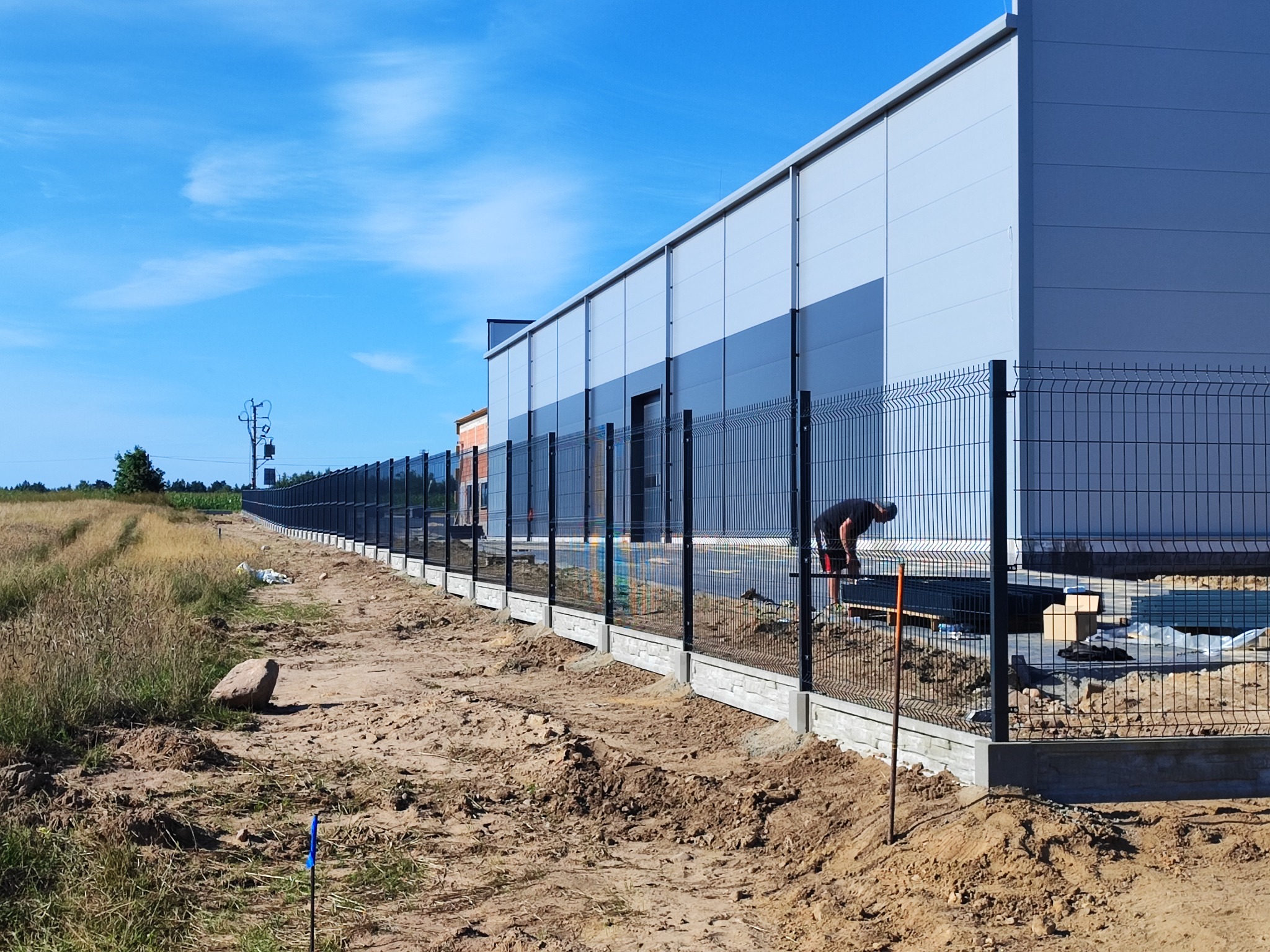 Długa linia nowoczesnego ogrodzenia panelowego z podmurówką, ciągnąca się wzdłuż budynku przemysłowego. Widoczny pracownik podczas montażu. Błękitne niebo.