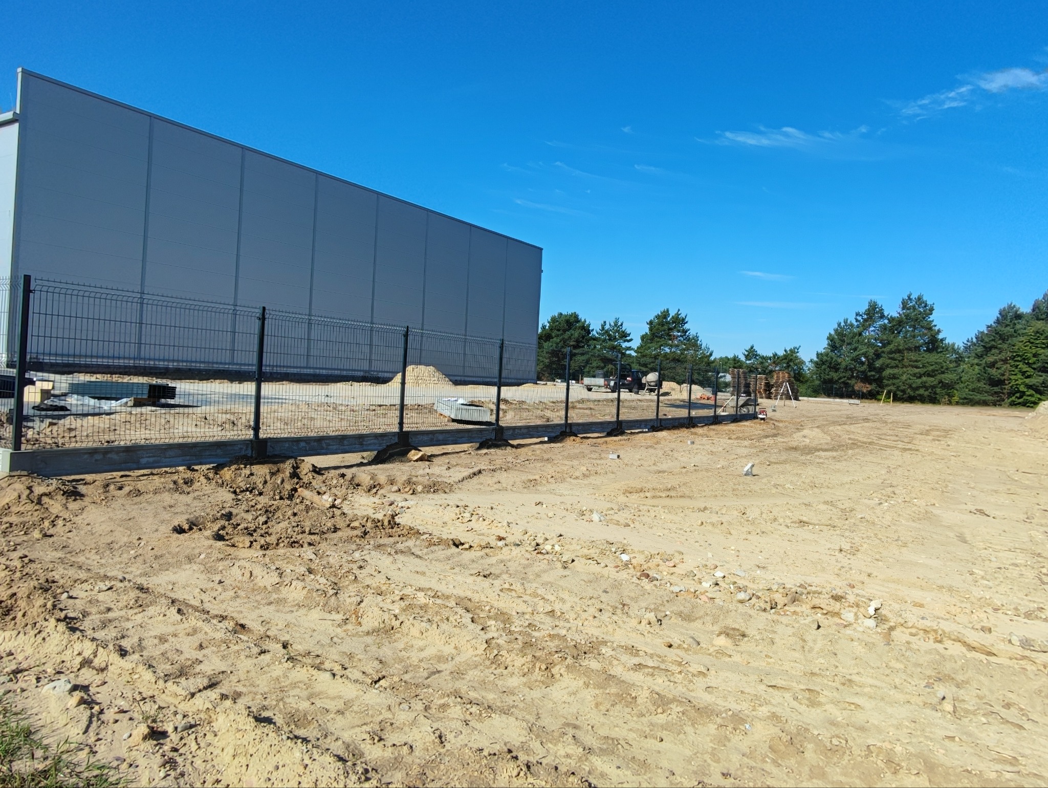 Częściowo zamontowane ogrodzenie panelowe na betonowej podmurówce, obok szarego budynku magazynowego, na piaszczystym terenie z widocznymi drzewami w tle, w słoneczny dzień.