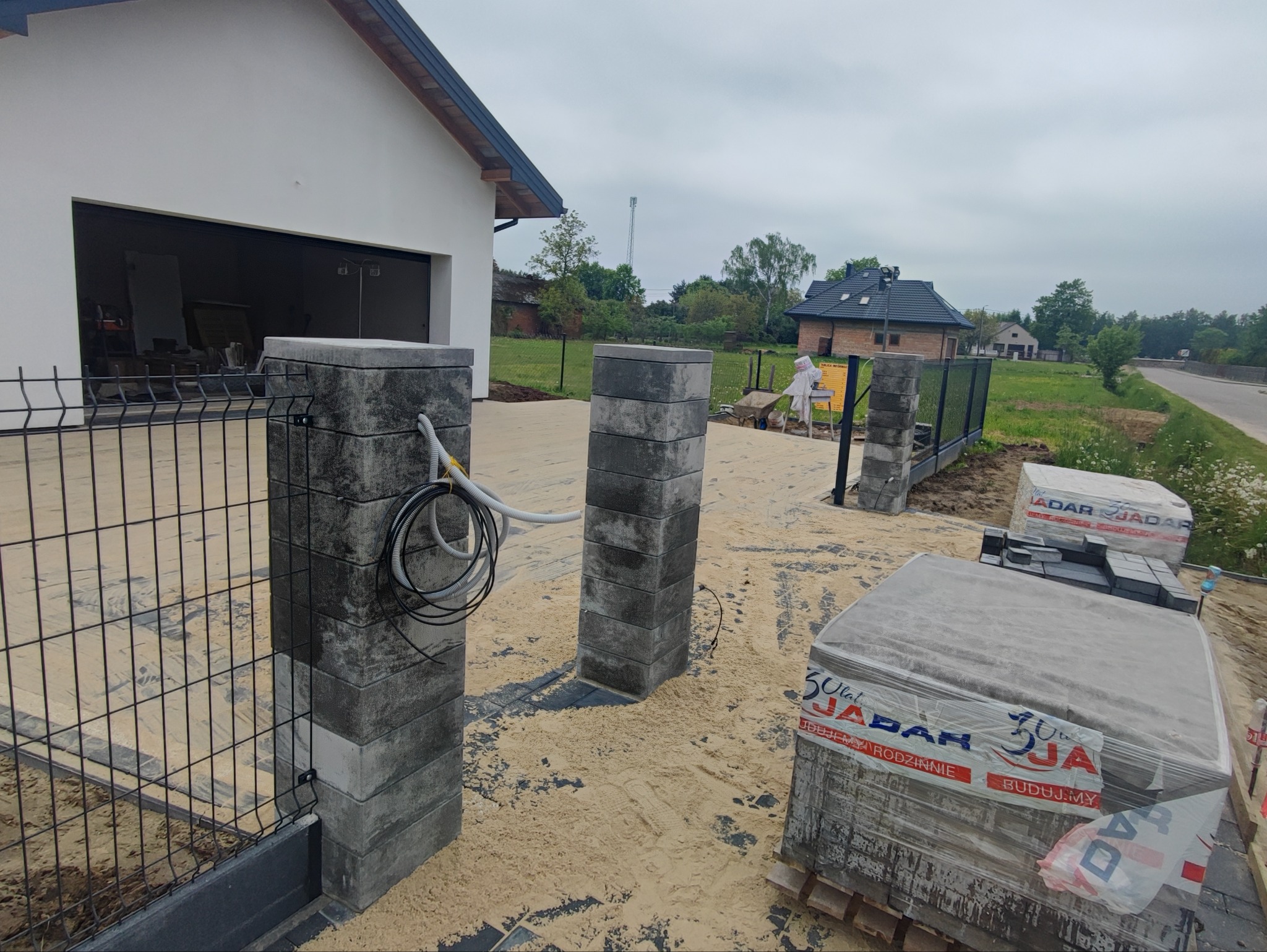 Montaż słupków ogrodzeniowych z szarej kostki betonowej, widoczne przewody elektryczne, częściowo zamontowane panele ogrodzeniowe i kostka brukowa na podjeździe.