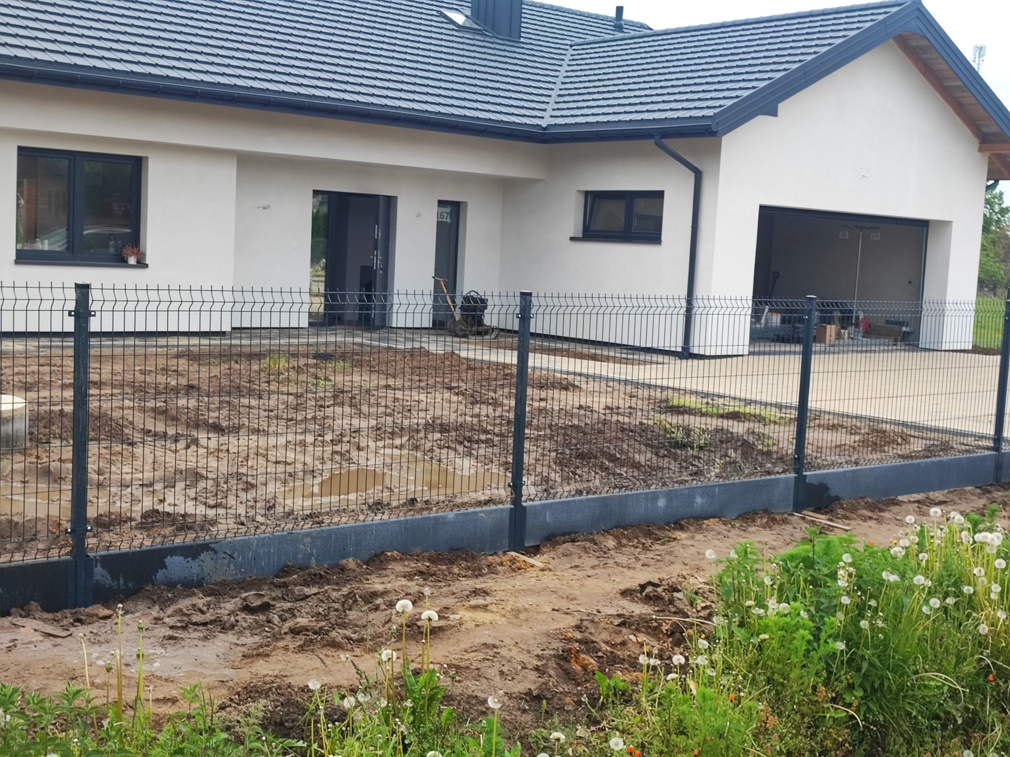 Nowoczesny dom z szarym ogrodzeniem panelowym na podmurówce. Widoczne świeżo przygotowane podłoże pod trawnik i wykończony podjazd z kostki.