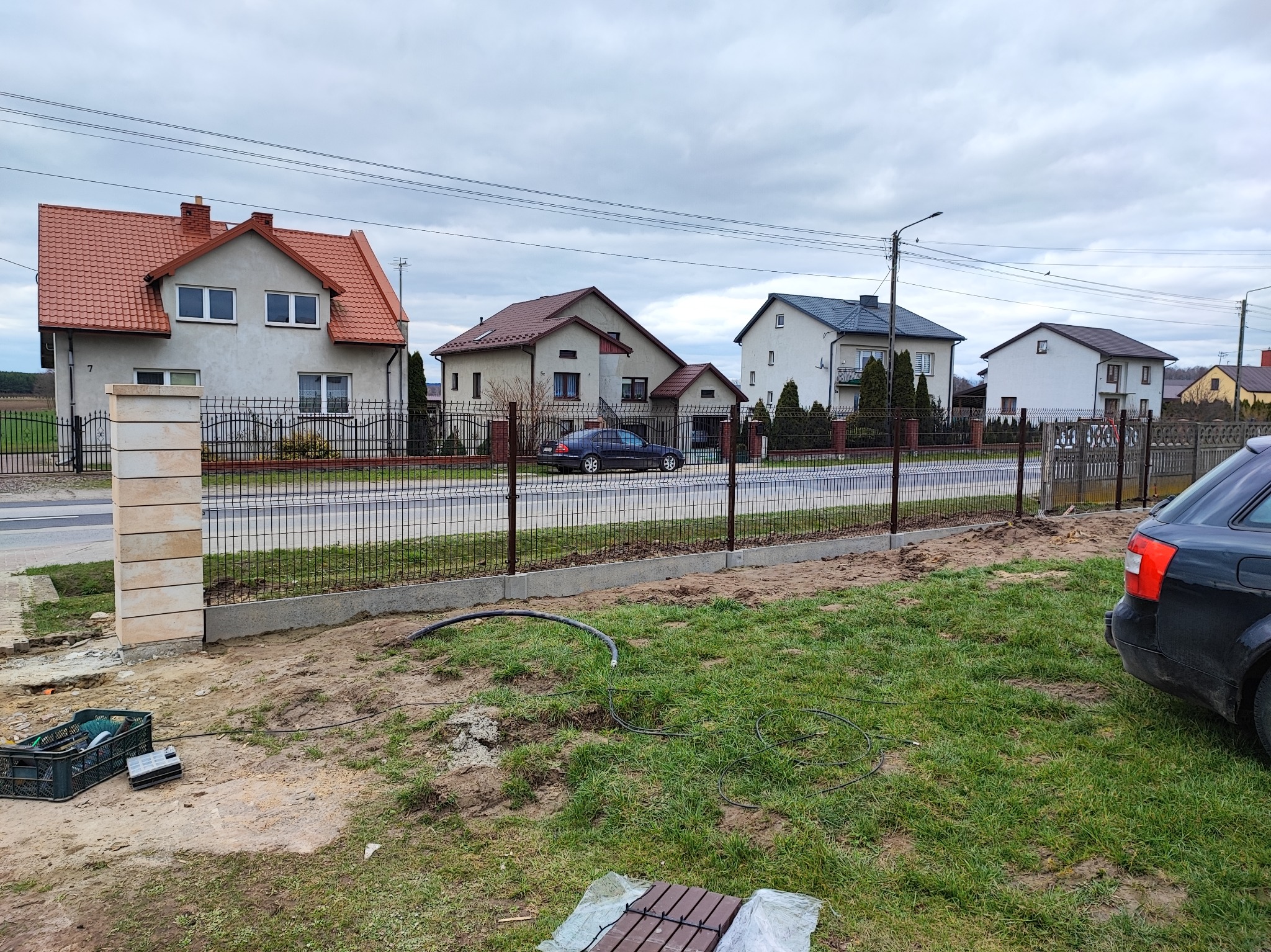 Wznoszenie ogrodzenia panelowego z podmurówką i słupkami z imitacji piaskowca. Widoczne narzędzia i materiały budowlane. Tło stanowią domy jednorodzinne.