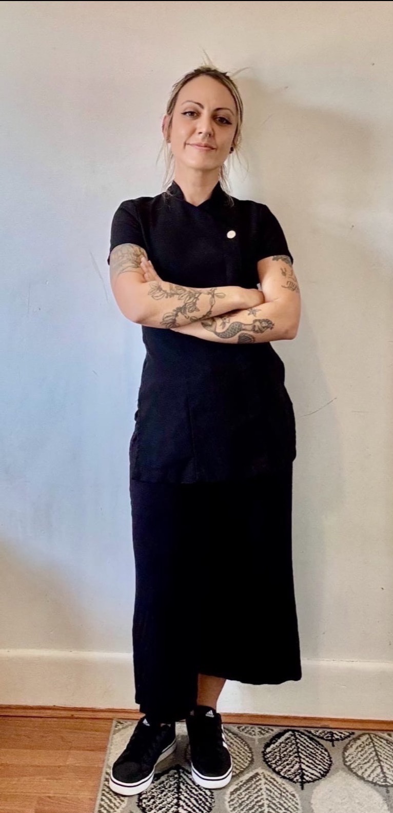 Masażystka w czarnym uniformie, z tatuażami na ramionach, stoi na tle jasnej ściany, prezentując profesjonalny wygląd i gotowość do pracy.