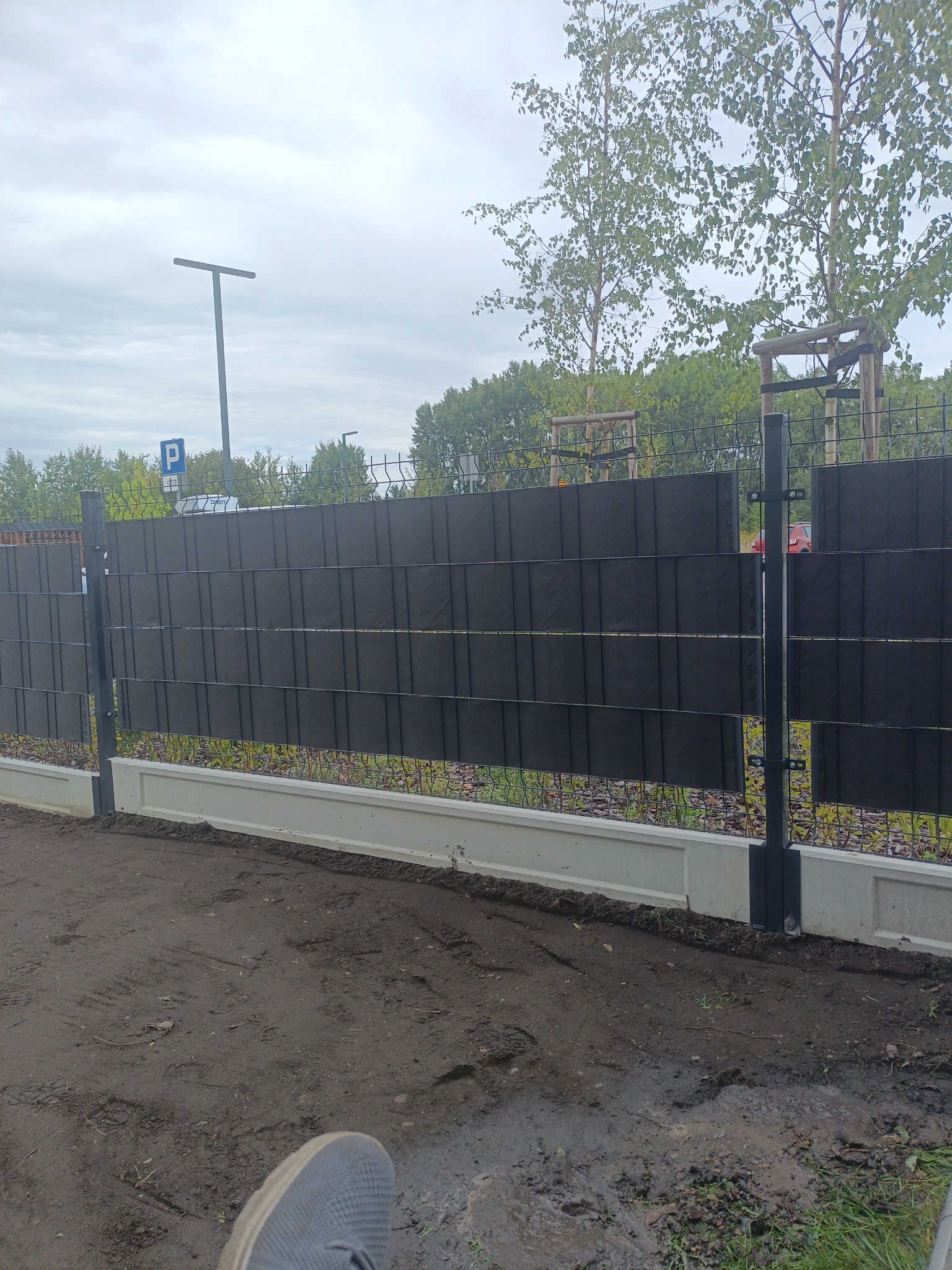 Nowoczesne ogrodzenie panelowe z podmurówką i czarnymi przesłonami, widoczne słupki i siatka. Tło: drzewa i parking. Ujęcie z dołu, fragment buta w kadrze.