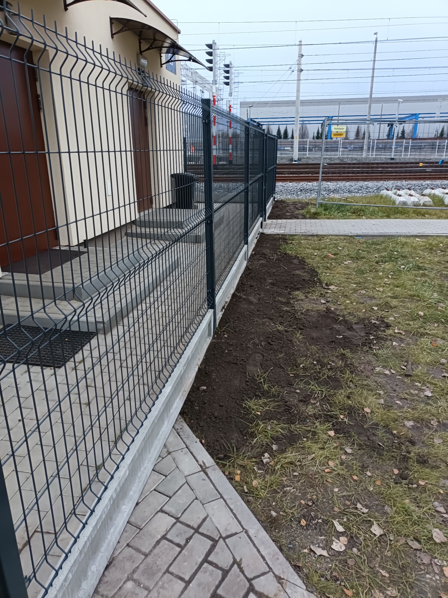 Nowoczesne ogrodzenie panelowe z podmurówką betonową, przylegające do budynku z kostką brukową. W tle widoczne tory kolejowe i budynek przemysłowy.