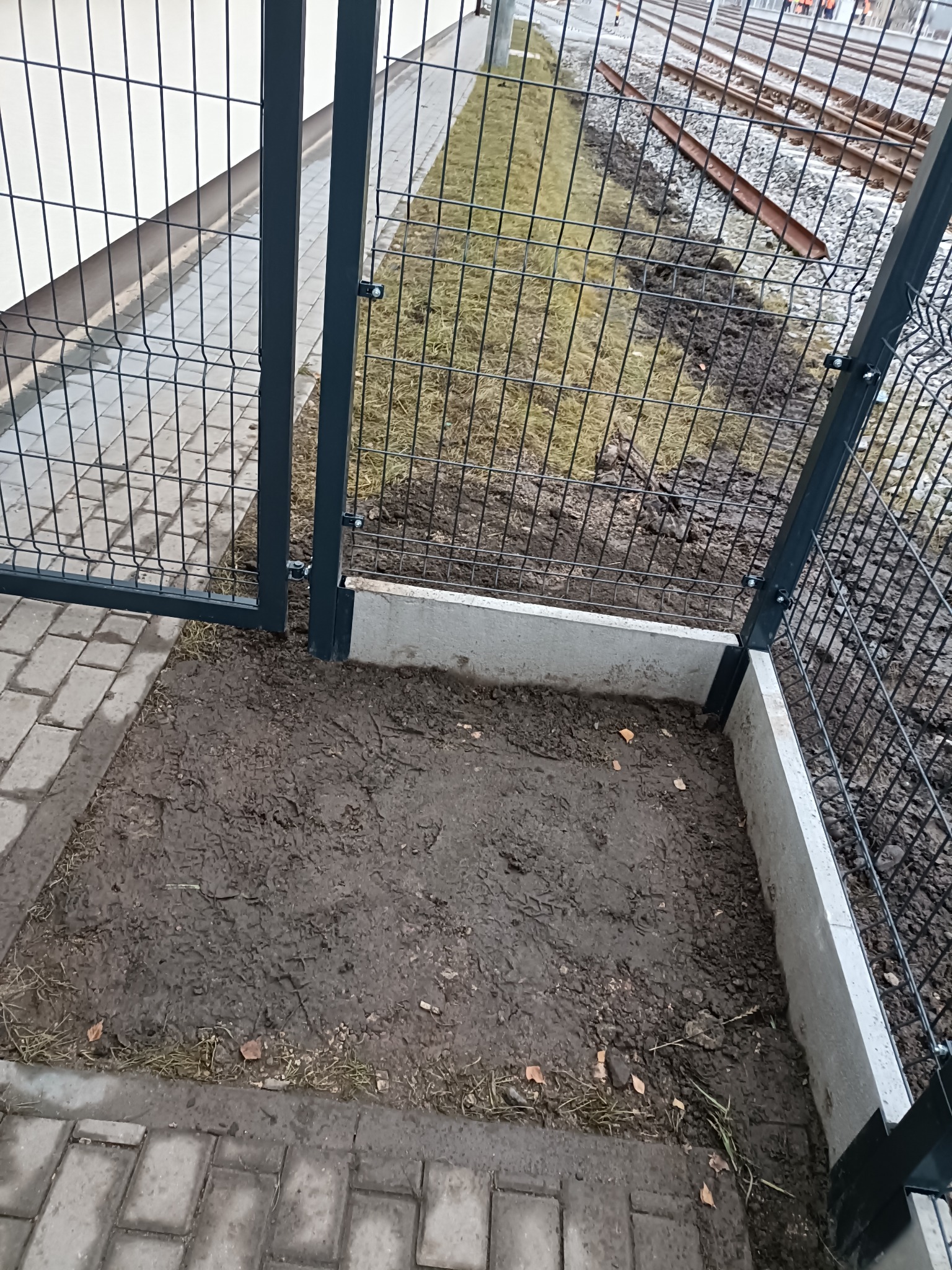 Fragment montażu ogrodzenia panelowego z podmurówką betonową, widok z góry na przygotowany grunt. W tle tory kolejowe.