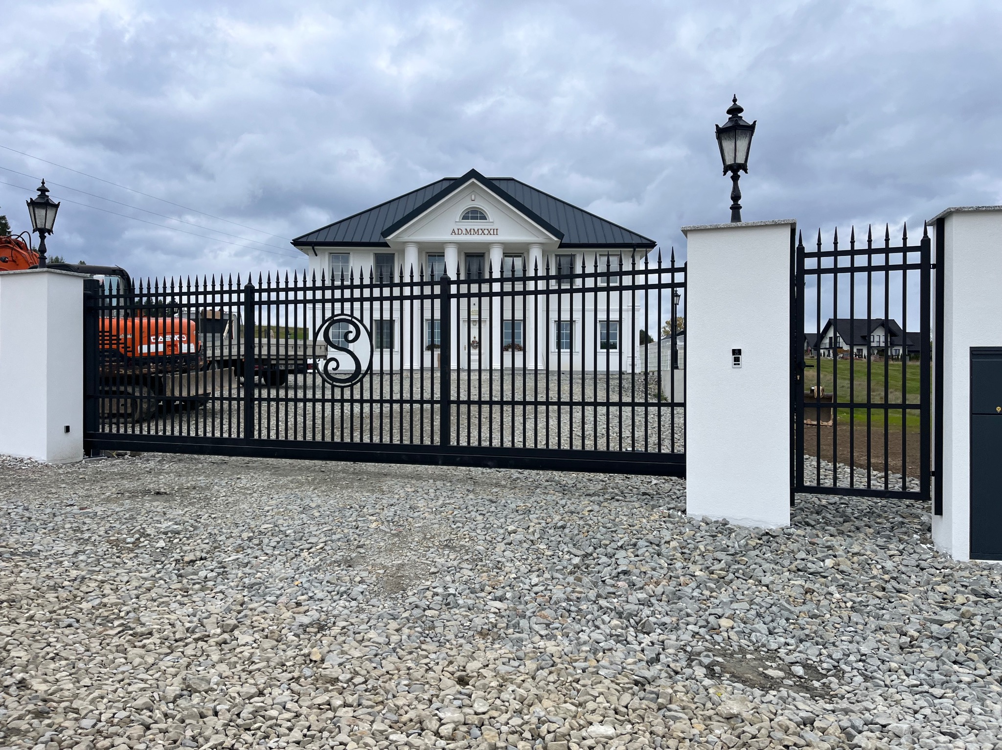 Czarna, kuta brama wjazdowa z inicjałem 'S' na tle białego, klasycznego domu z kolumnami i szarym niebem. Żwirowe podłoże.
