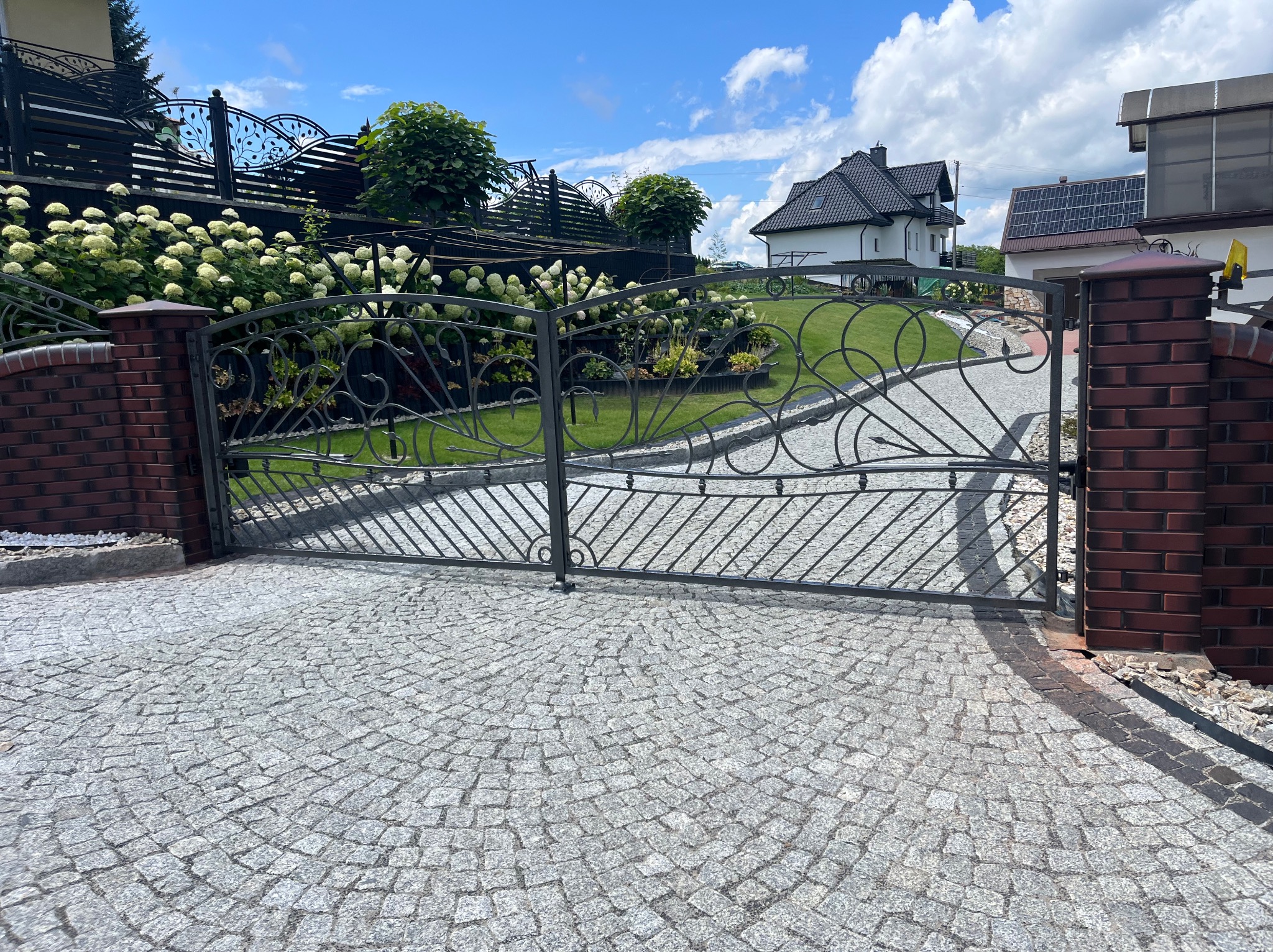 Ozdobna, metalowa brama wjazdowa z dekoracyjnymi elementami, osadzona na ceglanych słupkach. Tło stanowi zadbany ogród i dom z panelami słonecznymi.