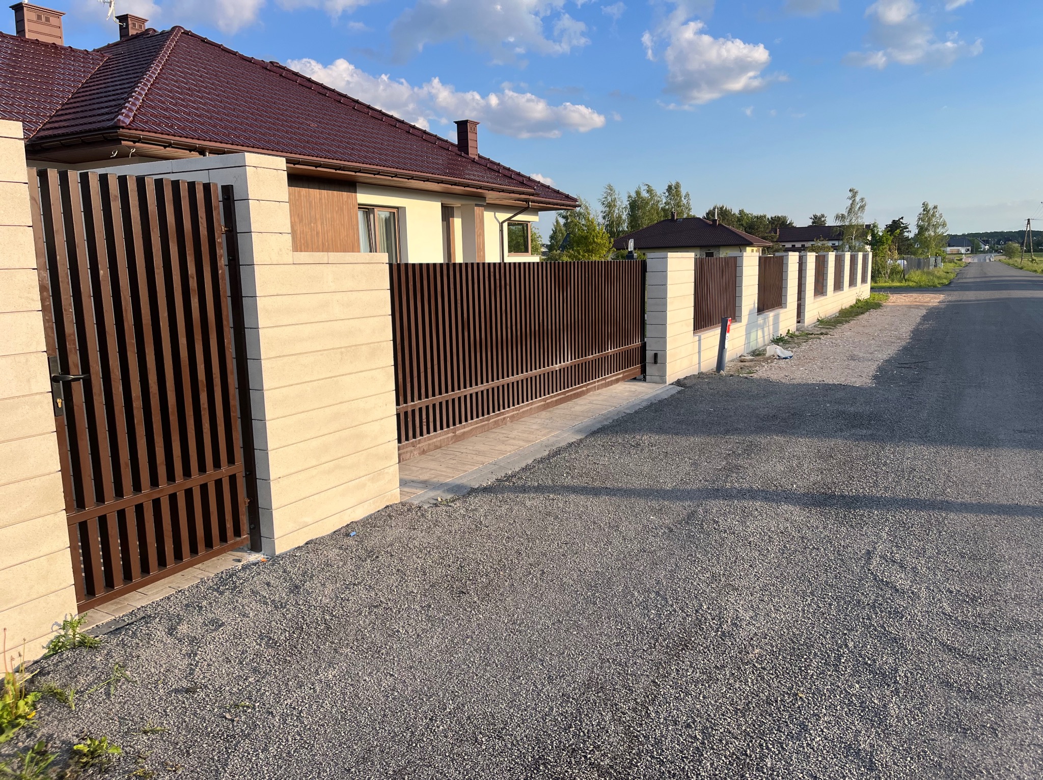 Nowoczesne ogrodzenie posesji z brązowymi, pionowymi panelami i jasnymi, blokowymi słupkami, widoczne z zewnątrz na tle domu z czerwoną dachówką i błękitnego nieba.