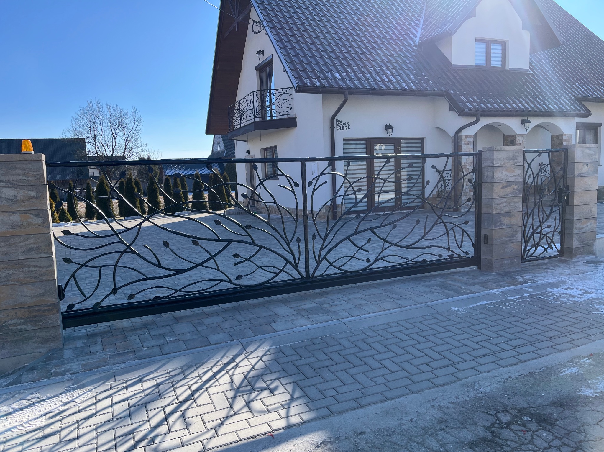 Ozdobna, przesuwna brama metalowa z motywem drzewa, zamontowana przed nowoczesnym domem z balkonem o podobnym wzorze. Brama z kamiennymi słupkami.