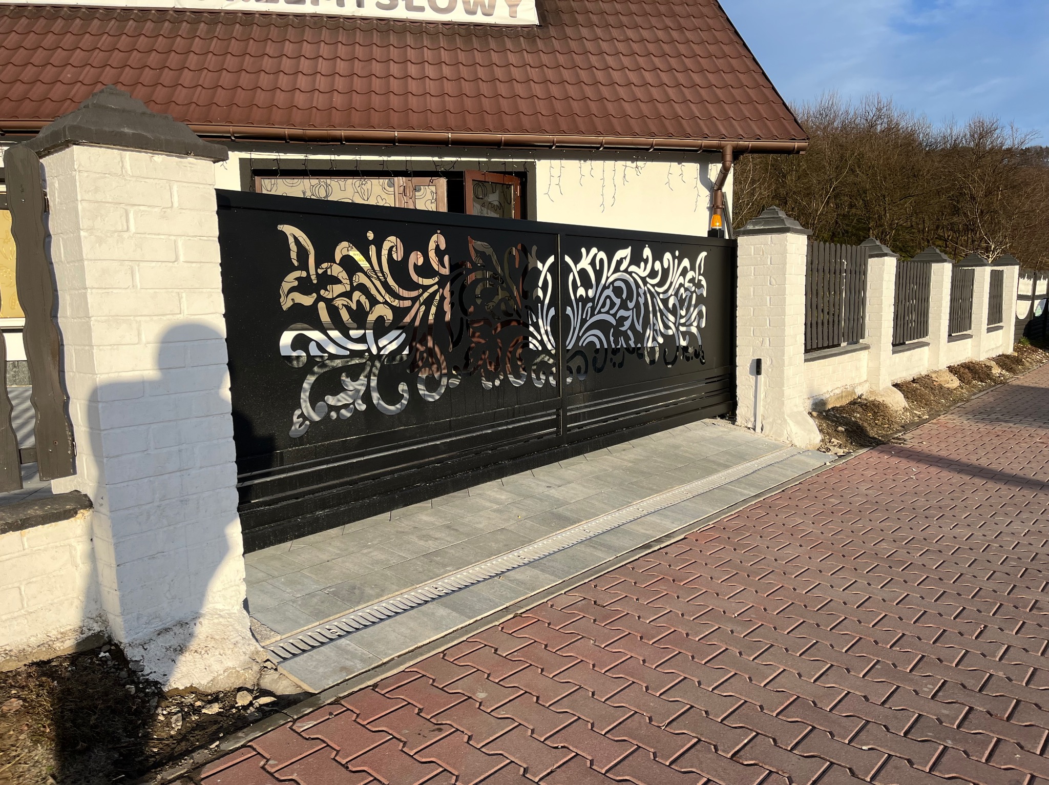 Czarna, metalowa brama wjazdowa z ażurowym, roślinnym wzorem, osadzona między murowanymi słupkami. W tle widoczny fragment ogrodzenia i budynek.