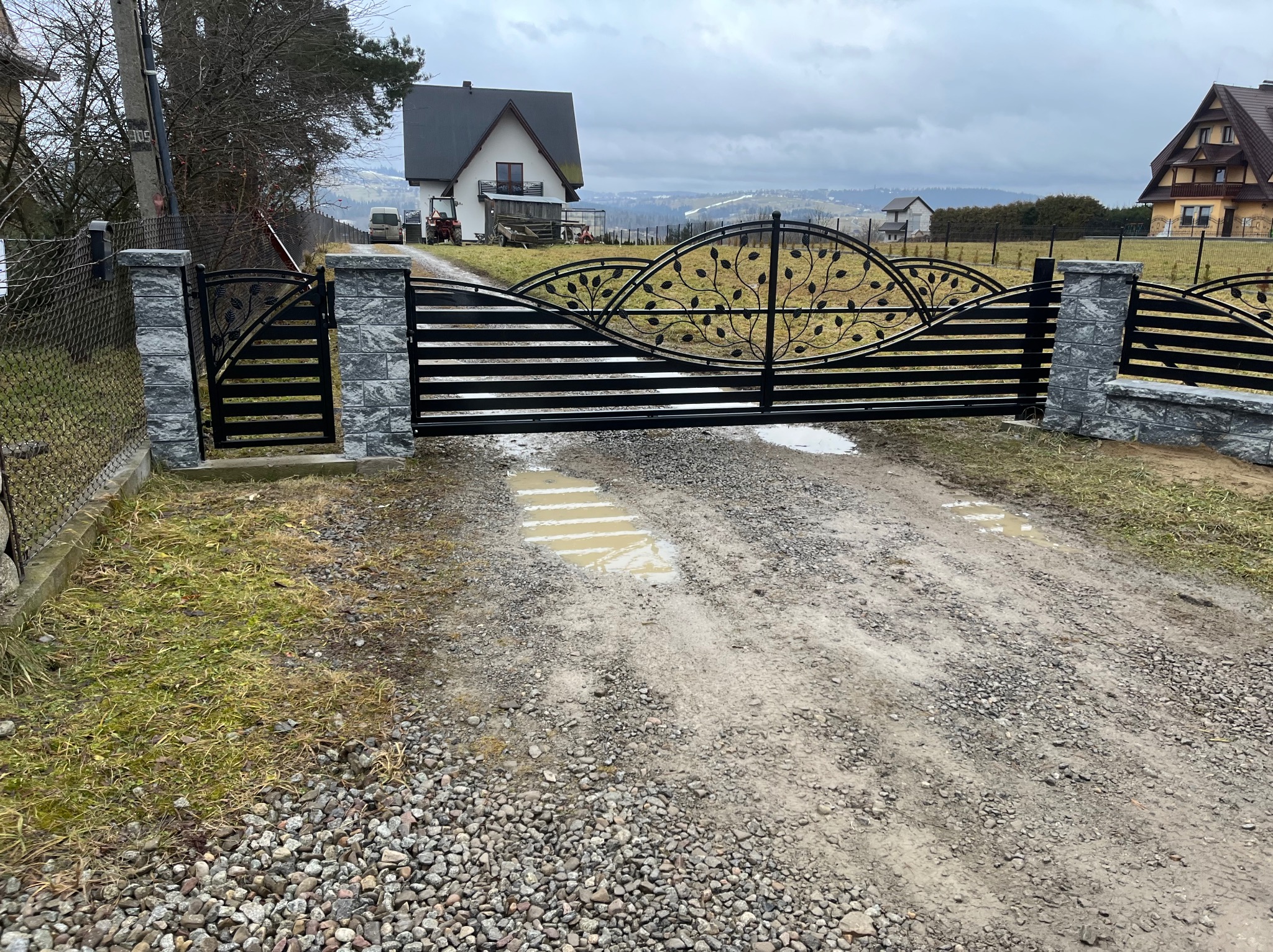 Czarna, metalowa brama wjazdowa z dekoracyjnym motywem liści, osadzona na kamiennych słupkach. W tle dom i pagórkowaty krajobraz.