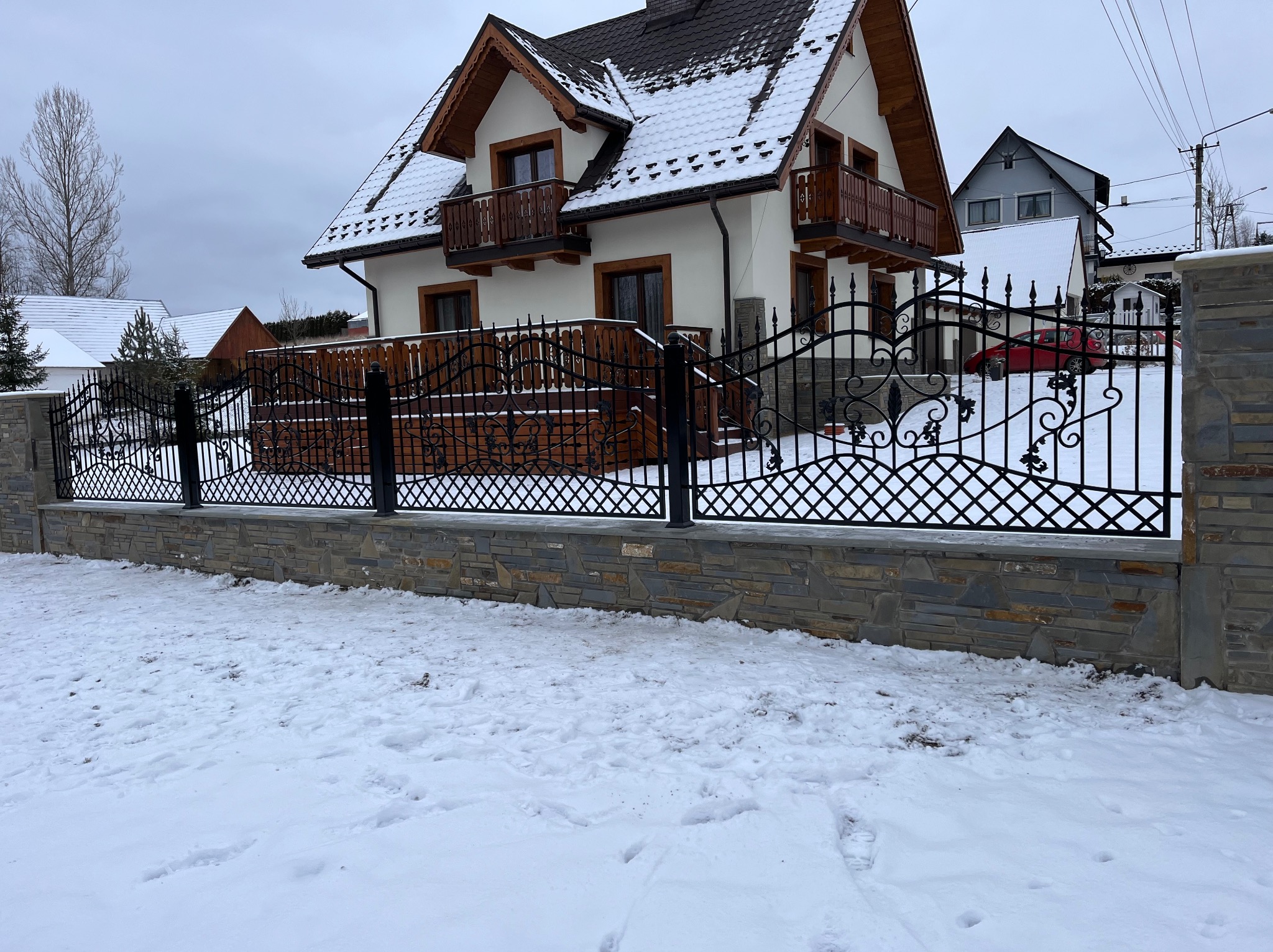 Kute ogrodzenie posesji z kamienną podmurówką i dekoracyjnymi elementami na tle ośnieżonego krajobrazu i domu z drewnianymi balkonami.