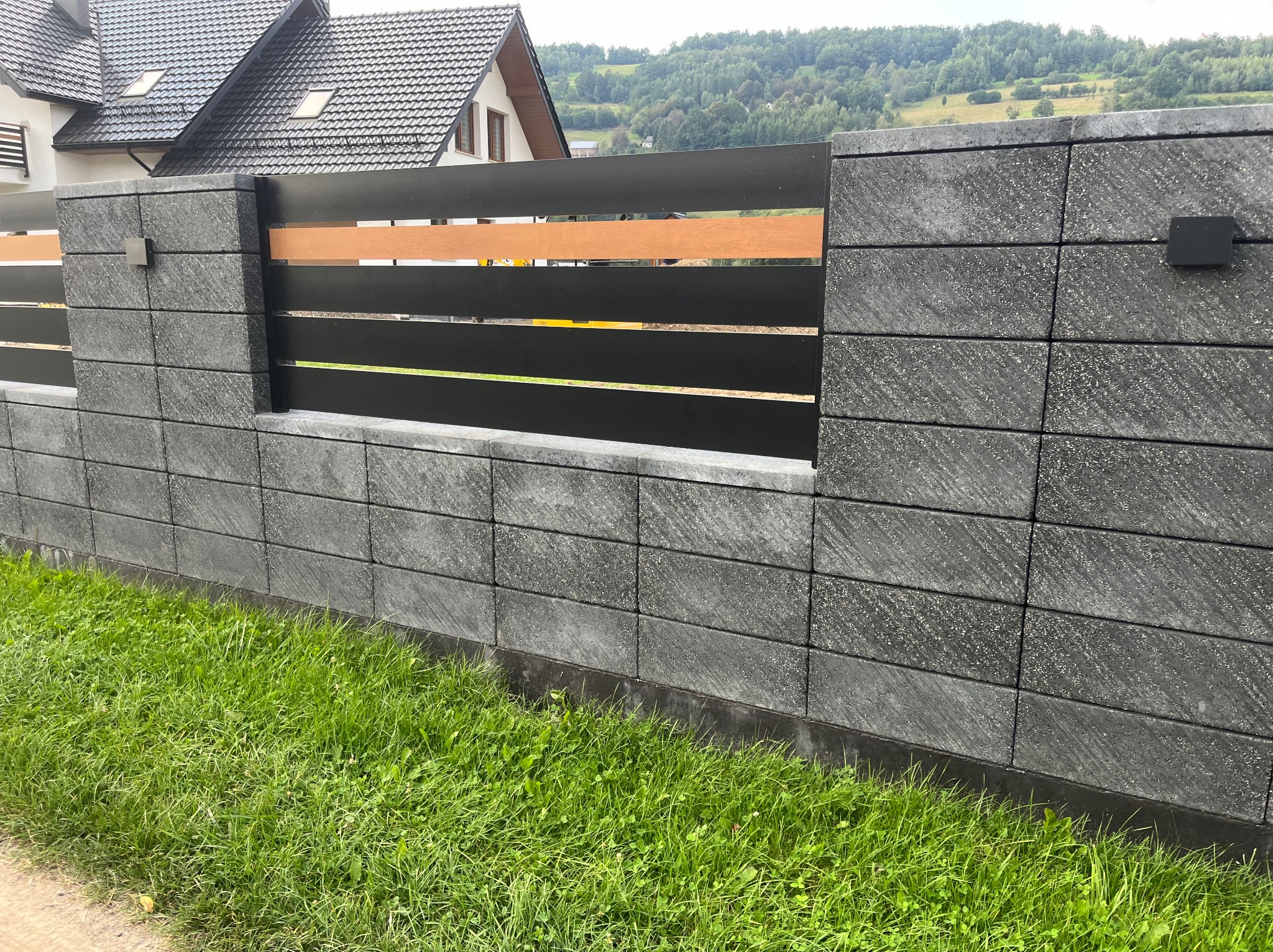 Nowoczesne ogrodzenie z betonowych bloczków i metalowych przęseł w Laskowej. Czarno-brązowa kolorystyka, widok na dom i zieleń w tle. Estetyczne wykończenie.
