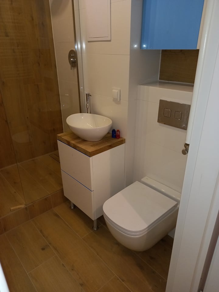 Nowoczesna łazienka z drewnopodobnymi płytkami, kabiną prysznicową typu walk-in i umywalką nablatową na szafce. Biała toaleta podwieszana i niebieska szafka wisząca.