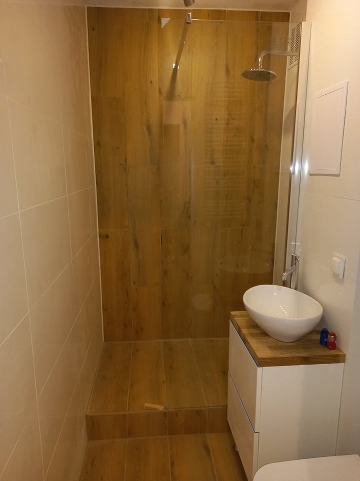 Nowoczesna kabina prysznicowa walk-in z drewnopodobnymi płytkami, umywalka nablatowa na białej szafce. Minimalistyczny design, jasne płytki na ścianach.