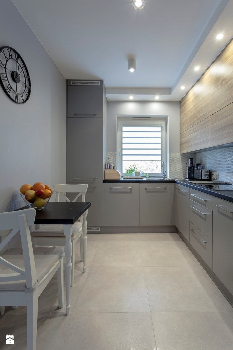 Nowoczesna kuchnia w szarościach z drewnianymi akcentami. Widoczny stół z owocami, zegar na ścianie i oświetlenie sufitowe.