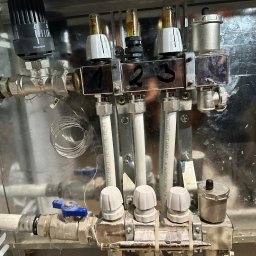 BARTŁOMIEJ MATERA SERWISNOW - Rozdzielacz ogrzewania podłogowego w metalowej szafce, z numerowanymi sekcjami (1-4), zaworami termostatycznymi i rurami z tworzywa sztucznego. Widoczne ślady użytkowania.