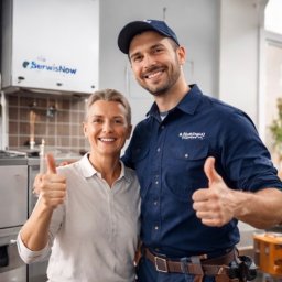 BARTŁOMIEJ MATERA SERWISNOW - Uśmiechnięty hydraulik w uniformie z klientką, oboje pokazują kciuki w górę na tle pieca gazowego 'SerwisNow' i płytek. Dobre relacje z klientem.