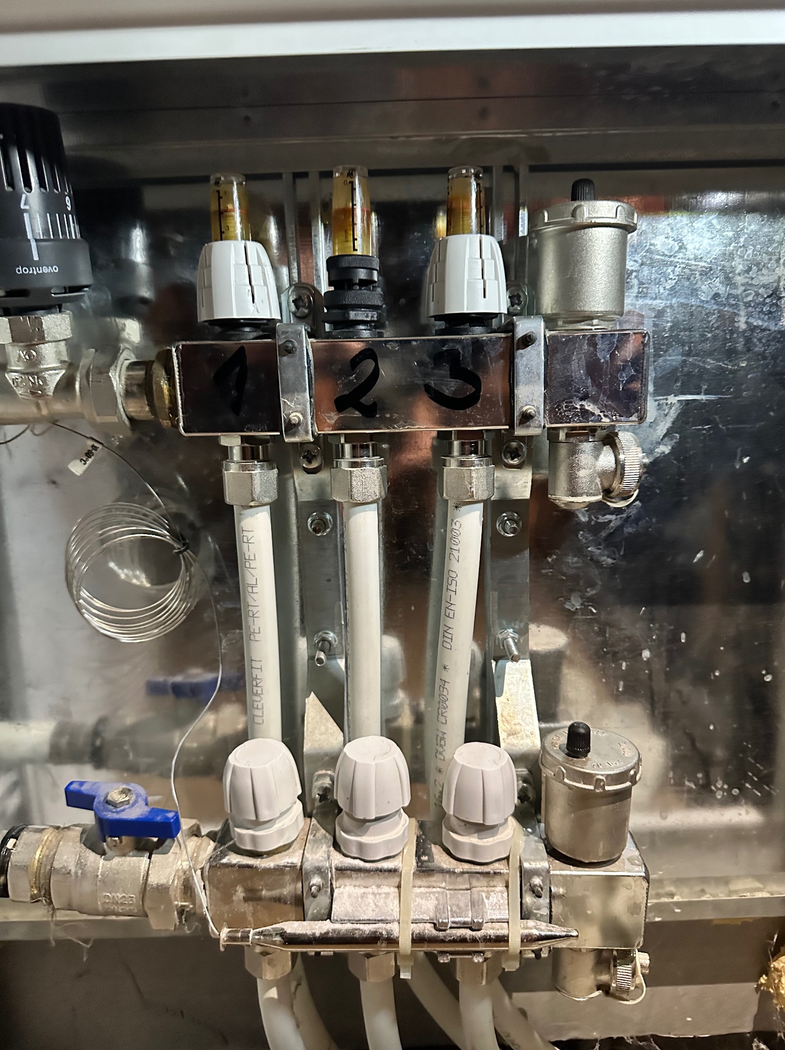 Rozdzielacz instalacji grzewczej z numeracją 1, 2, 3, zaworami i rurami z tworzywa, zamontowany w metalowej obudowie. Widoczne elementy regulacyjne i przyłączeniowe.