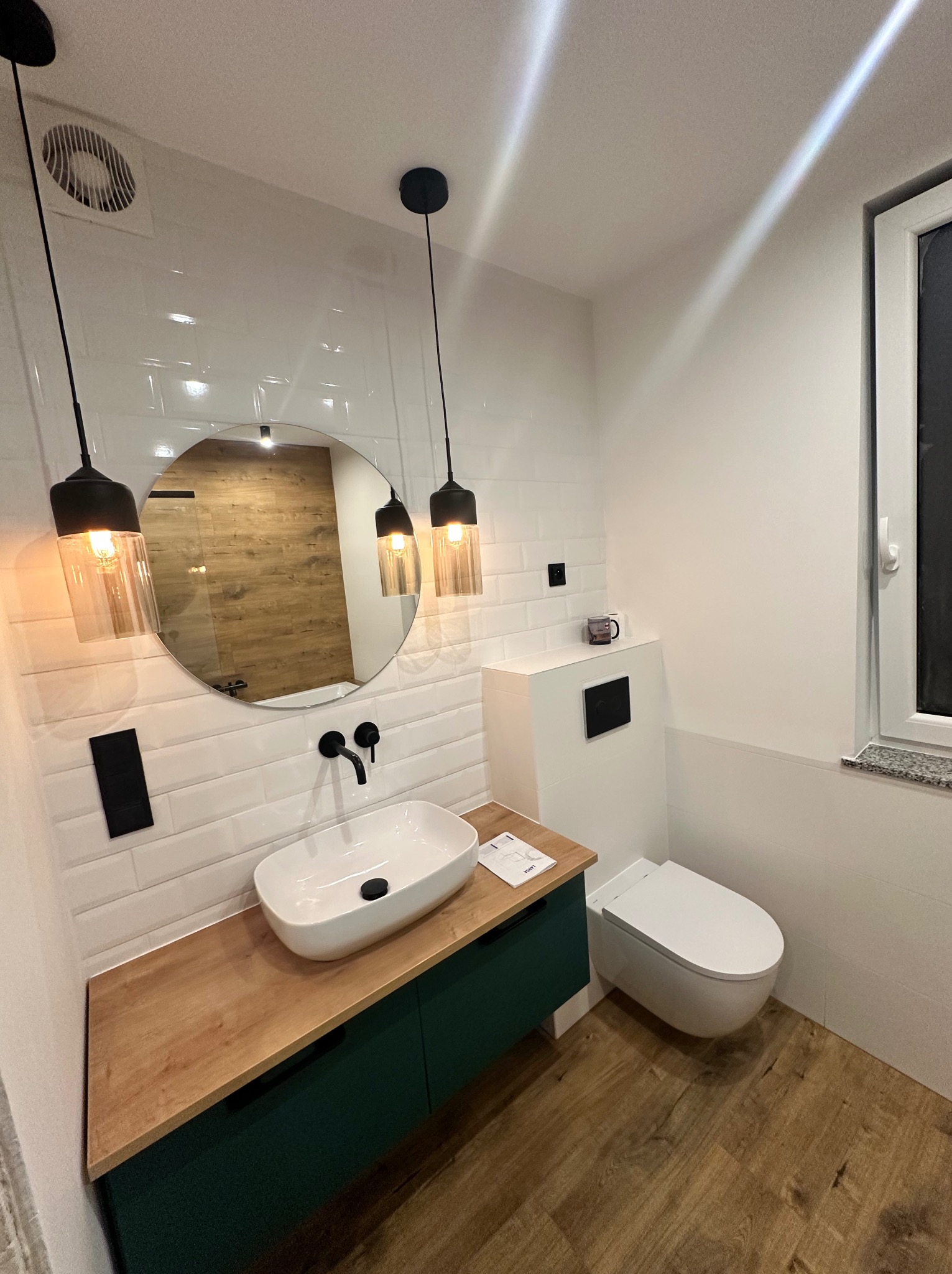 Nowoczesna łazienka z owalną umywalką, drewnianym blatem i zieloną szafką. Na ścianie białe płytki, okrągłe lustro i wiszące lampy. Widoczna toaleta i drewnopodobna podłoga.