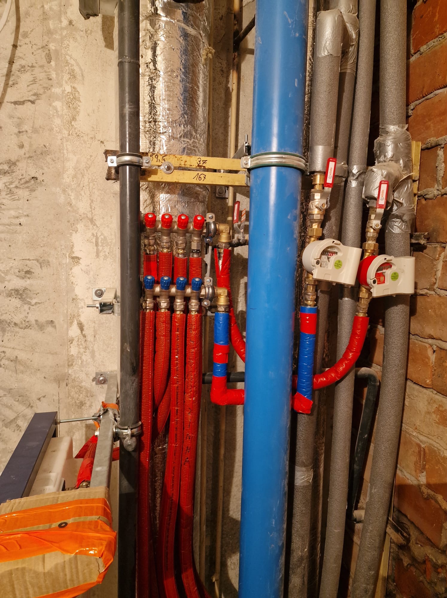 Rozdzielacz ogrzewania podłogowego z czerwonymi rurami, niebieska rura pionowa, szare rury izolowane, liczniki ciepła i zawory na tle surowej ściany.