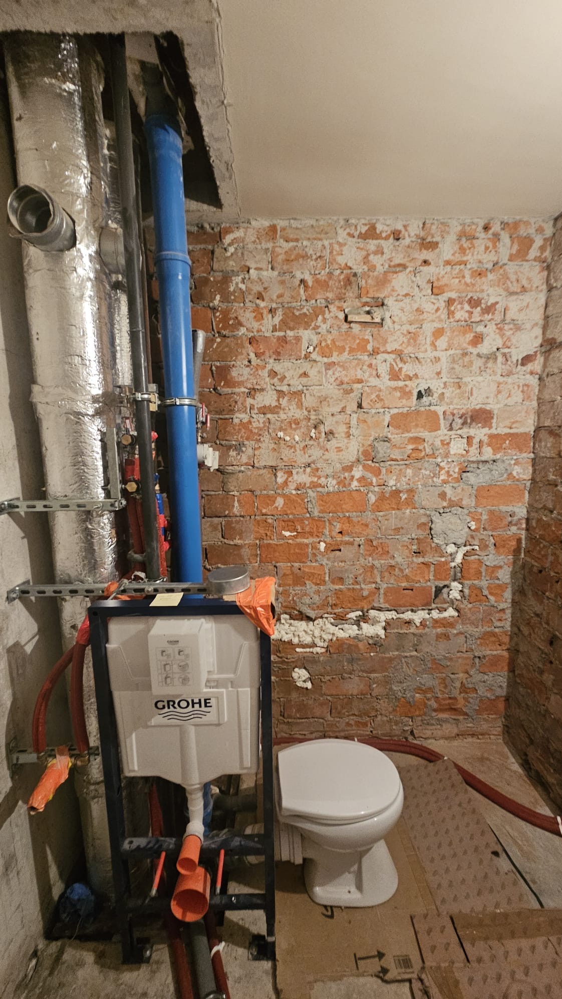 Montaż stelaża podtynkowego Grohe z widocznymi rurami instalacyjnymi na tle surowej, ceglanej ściany. Częściowo zamontowana toaleta. Prace hydrauliczne w trakcie realizacji.