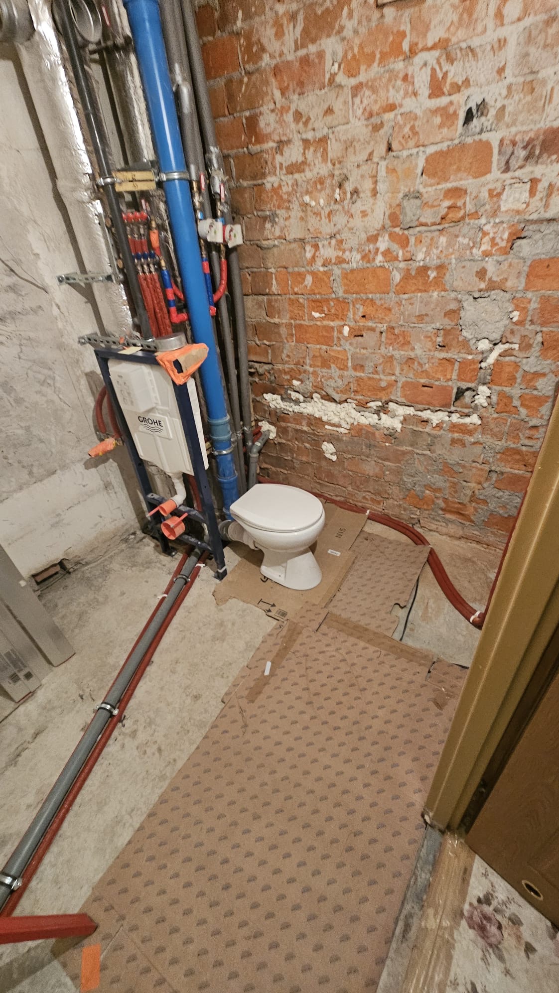 Biała toaleta z podtynkową spłuczką Grohe na tle surowej ceglanej ściany i instalacji hydraulicznej. Podłoga zabezpieczona kartonem.