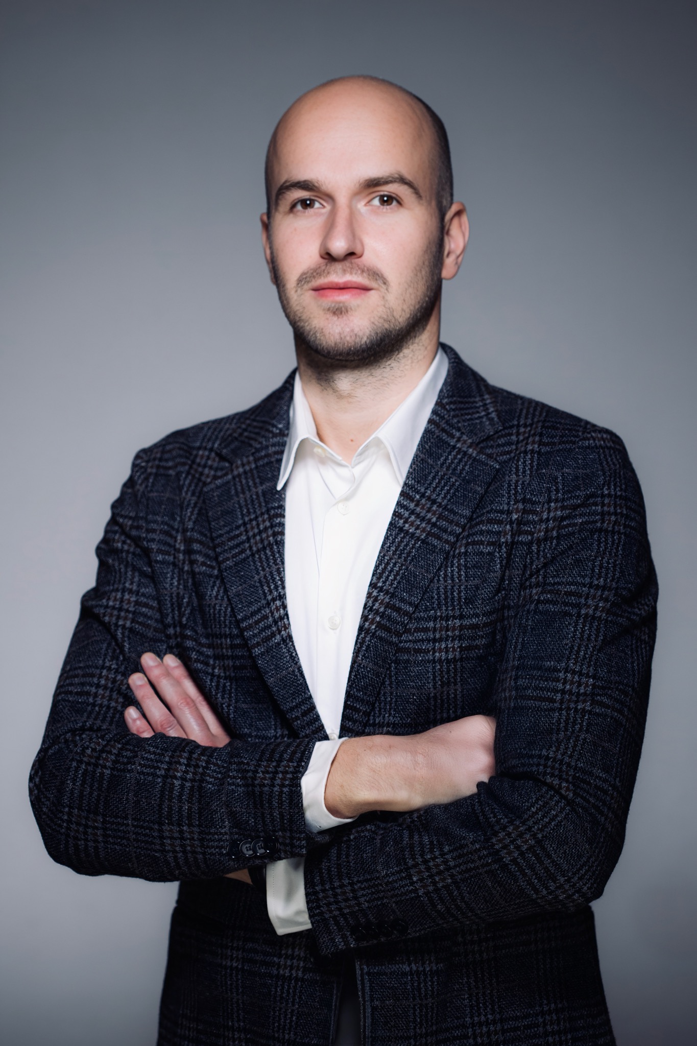 Portret mężczyzny w garniturze w kratę i białej koszuli, z założonymi rękami na szarym tle. Profesjonalny wizerunek.