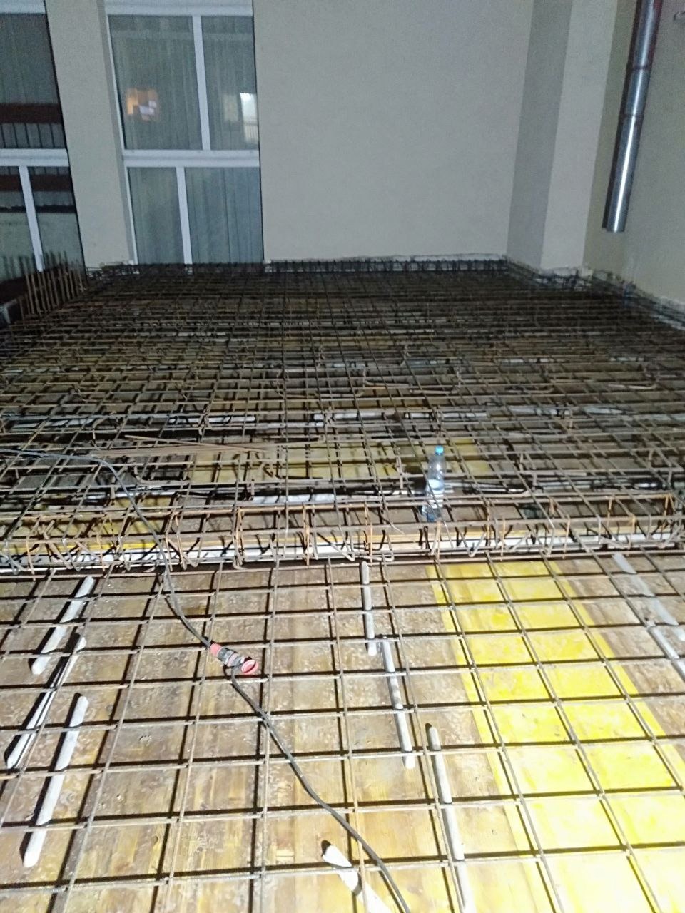 Zbrojenie stropu na żółtym szalunku, widoczne pręty zbrojeniowe ułożone w kratę, butelka wody i przewód elektryczny. W tle ściana i okno.