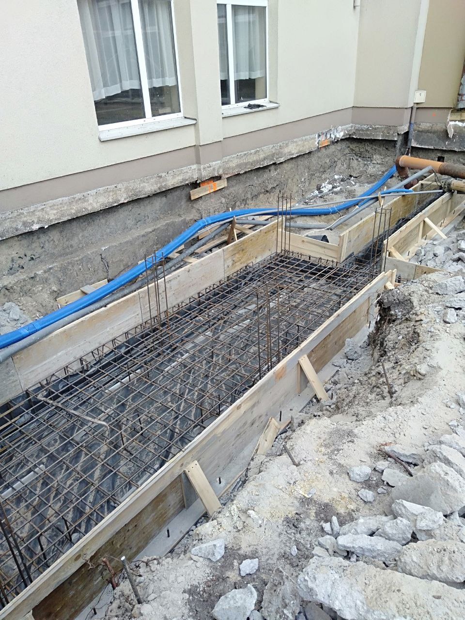 Zbrojenie fundamentu w szalunku drewnianym, widoczne rury instalacyjne i fragment budynku w tle. Detal zbrojenia siatką stalową przed betonowaniem.