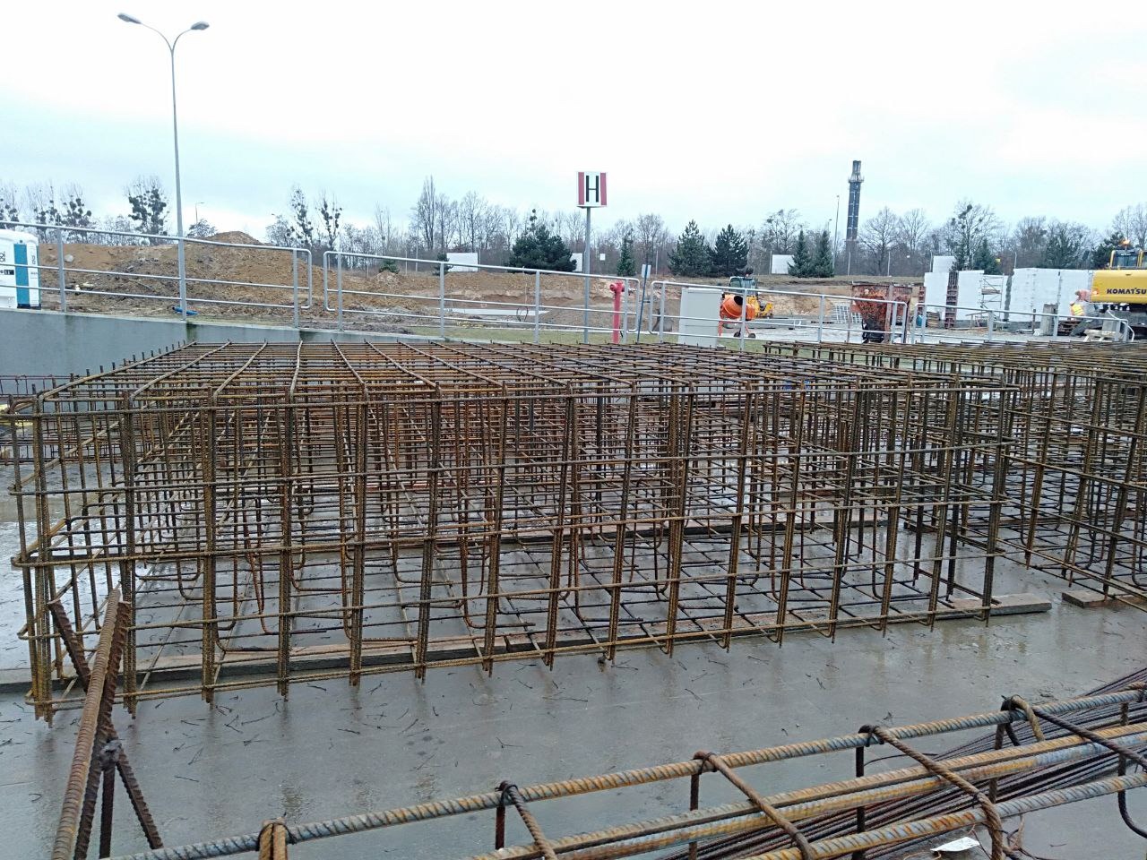 Siatka zbrojeniowa w formie prostopadłościanu, przygotowana do zalania betonem na budowie. Widoczne elementy infrastruktury i sprzęt budowlany w tle.