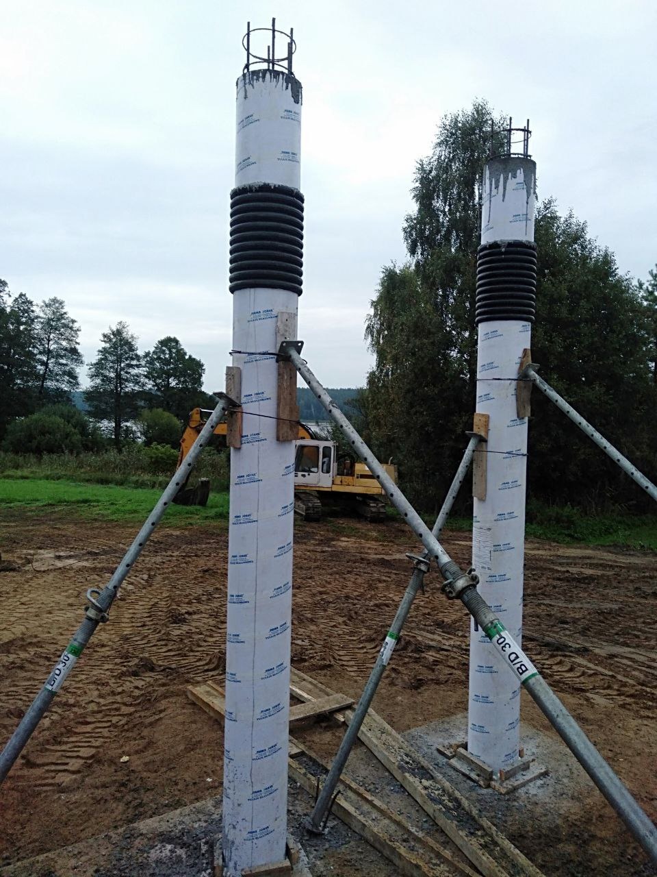 Dwa filary szalunkowe zbrojone, wsparte na podporach, na placu budowy z koparką w tle. Widoczne ślady opon na ziemi i elementy drewniane szalunku.