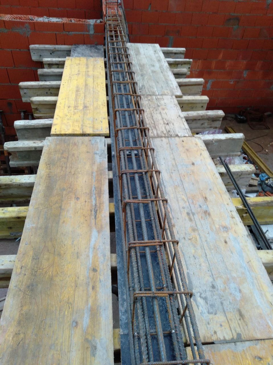 Zbrojenie pionowe w szalunku, przygotowane do zalania betonem. Widoczne deski szalunkowe i ceglany mur w tle. Detal z budowy budynku żelbetowego.